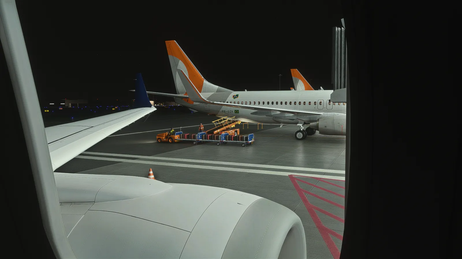 View from a cabin window over the wing: a neighbouring 737 with baggage carts and a belt loader at night.